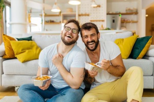 A male couple eating pasta out of takeout co<em></em>ntainers while sitting on their living room floor and laughing.